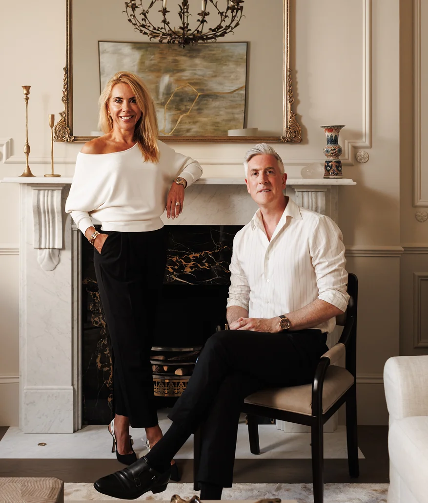 Two interior designers posed in an elegant living room with a fireplace and classic decor.