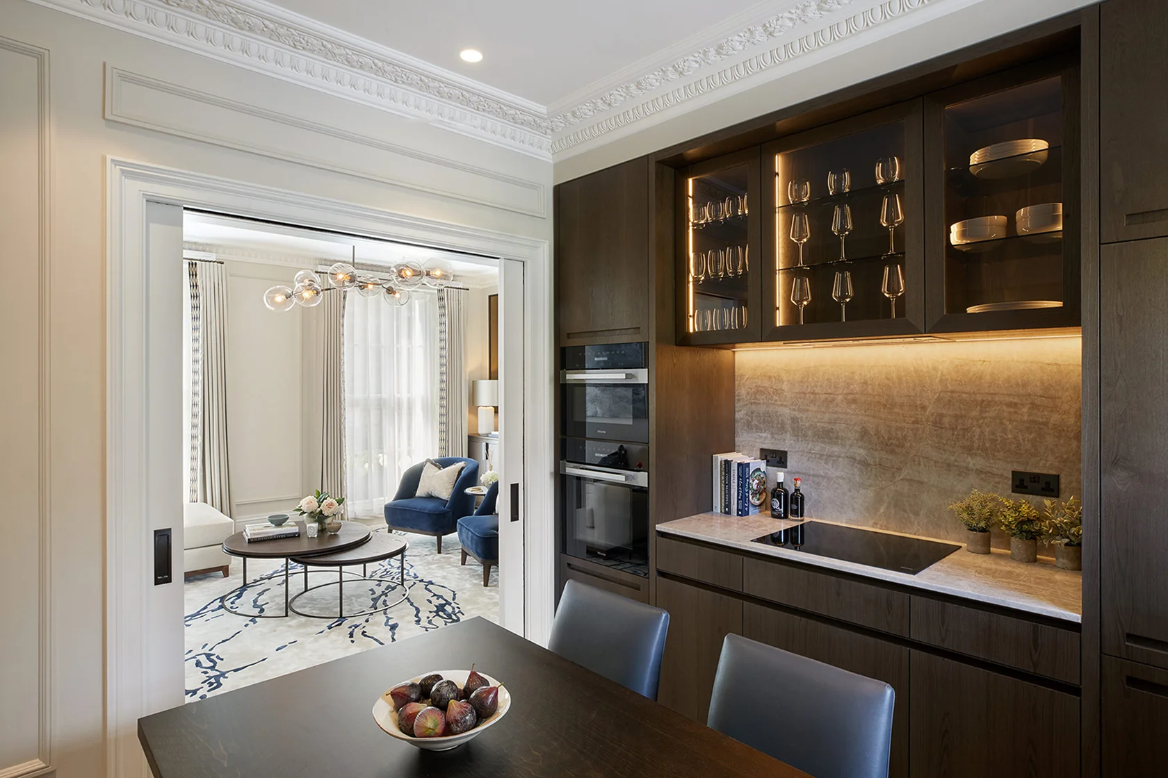 Modern kitchen with dark cabinets, illuminated countertop, and a dining table with a fruit bowl, connected to a bright