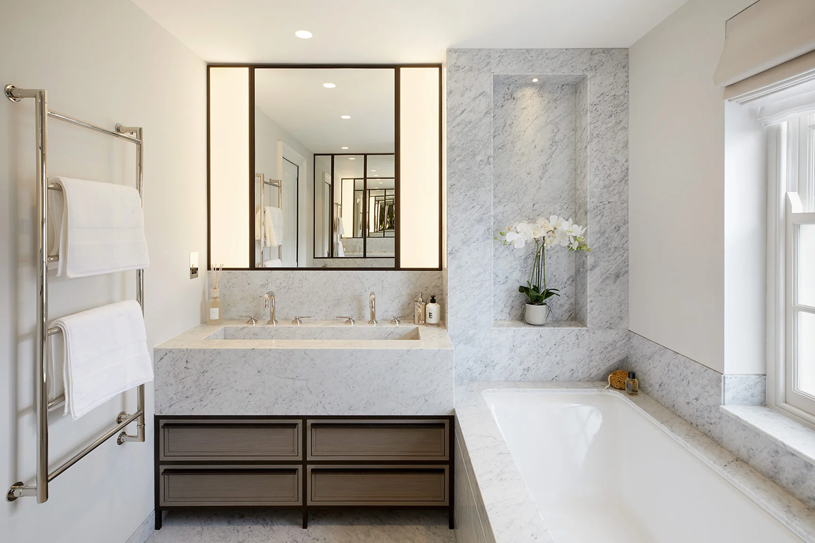 Modern bathroom featuring a marble vanity, bathtub, large mirror, and potted white orchids. Soft lighting illuminates the