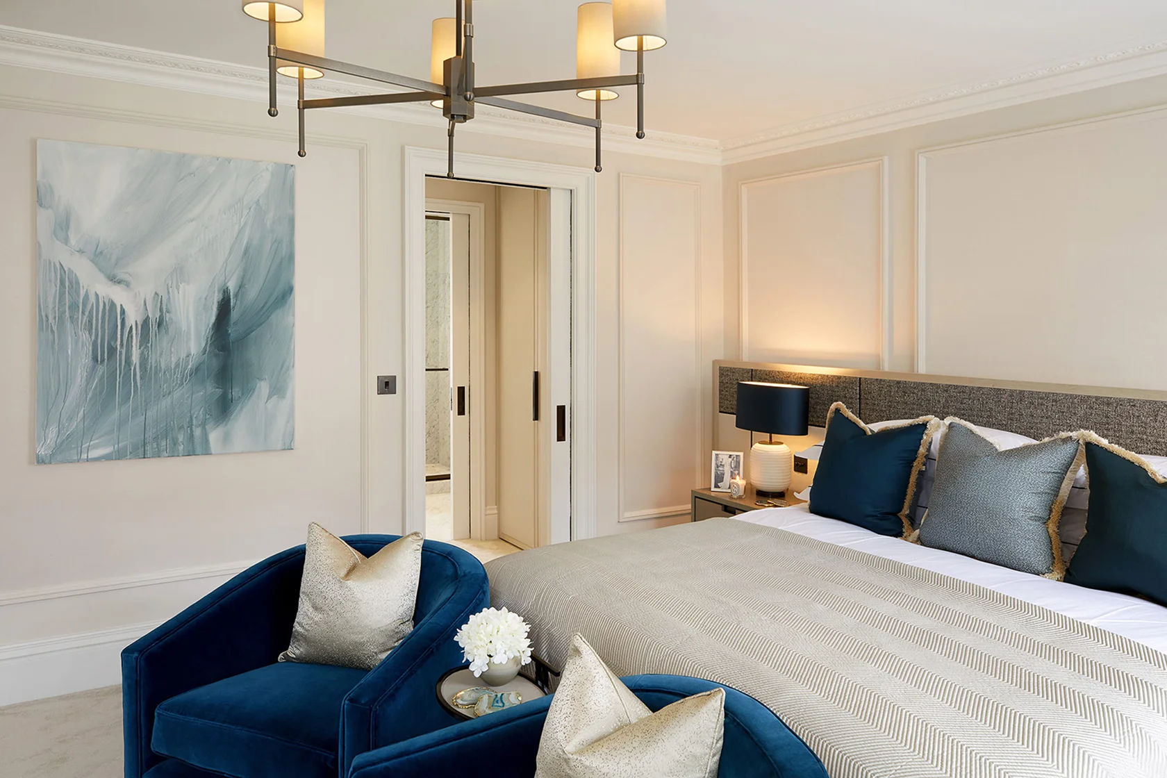 Luxurious bedroom featuring a large bed, blue accent chairs, modern lighting, and abstract artwork on the wall.