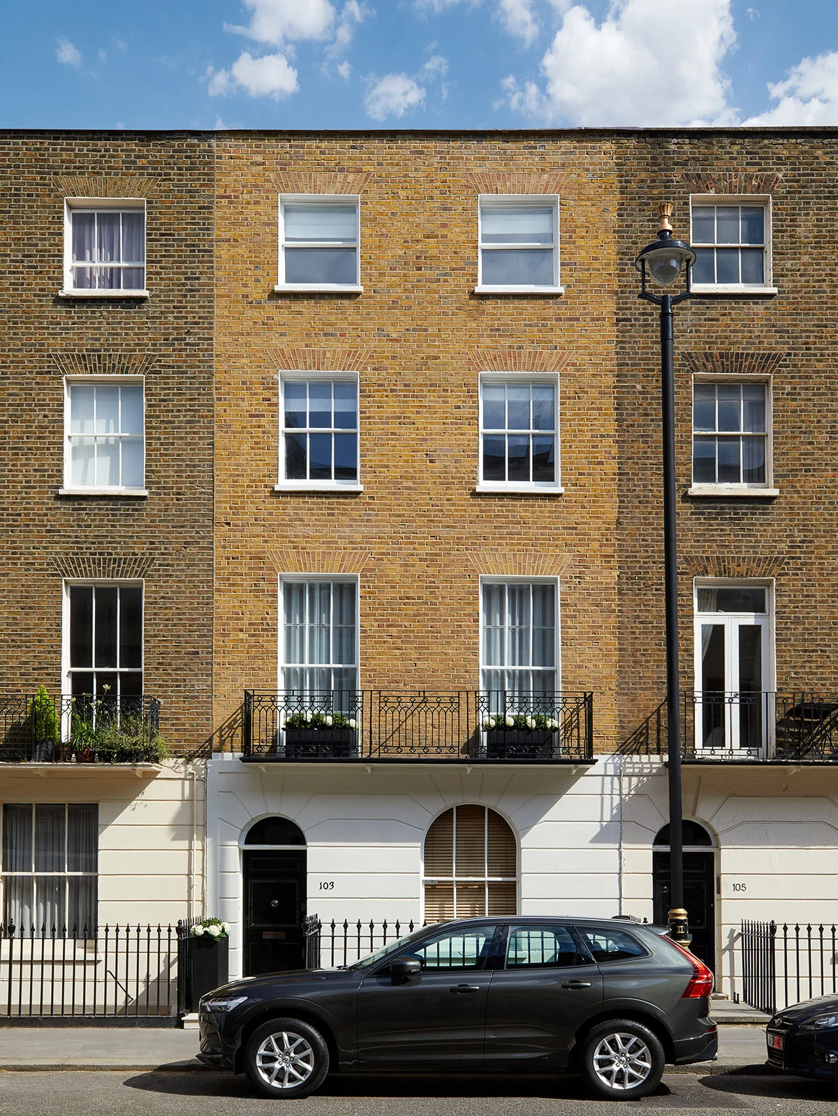 Three-story brick building with white window frames and balcony, parked car in front, blue sky with clouds above.