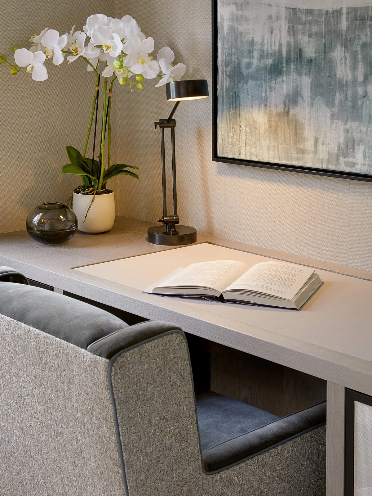 A modern workspace featuring a desk, open book, lamp, and potted orchids, with abstract wall art in the background.