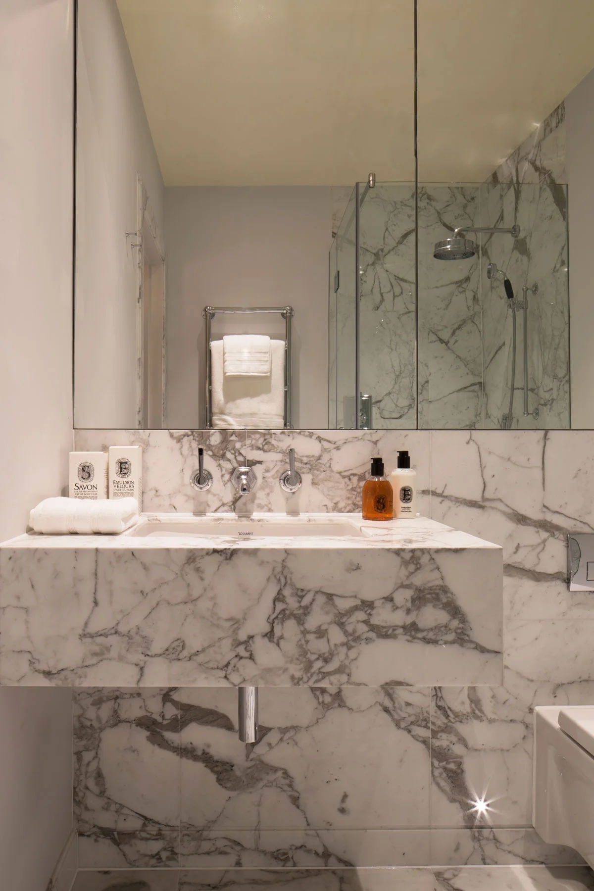 Modern bathroom with marble countertop, sink, and large mirror, featuring toiletries and a glass shower enclosure.