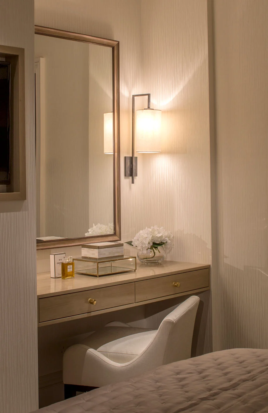 Cozy vanity area with a mirror, a lamp, books, and a white chair, illuminated with soft lighting.