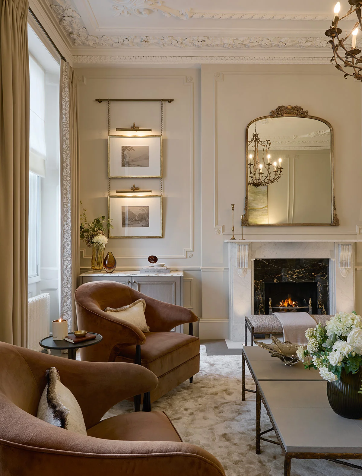 Elegant living room with two brown armchairs, a fireplace, decorative mirror, and soft lighting. Floral arrangement on table.