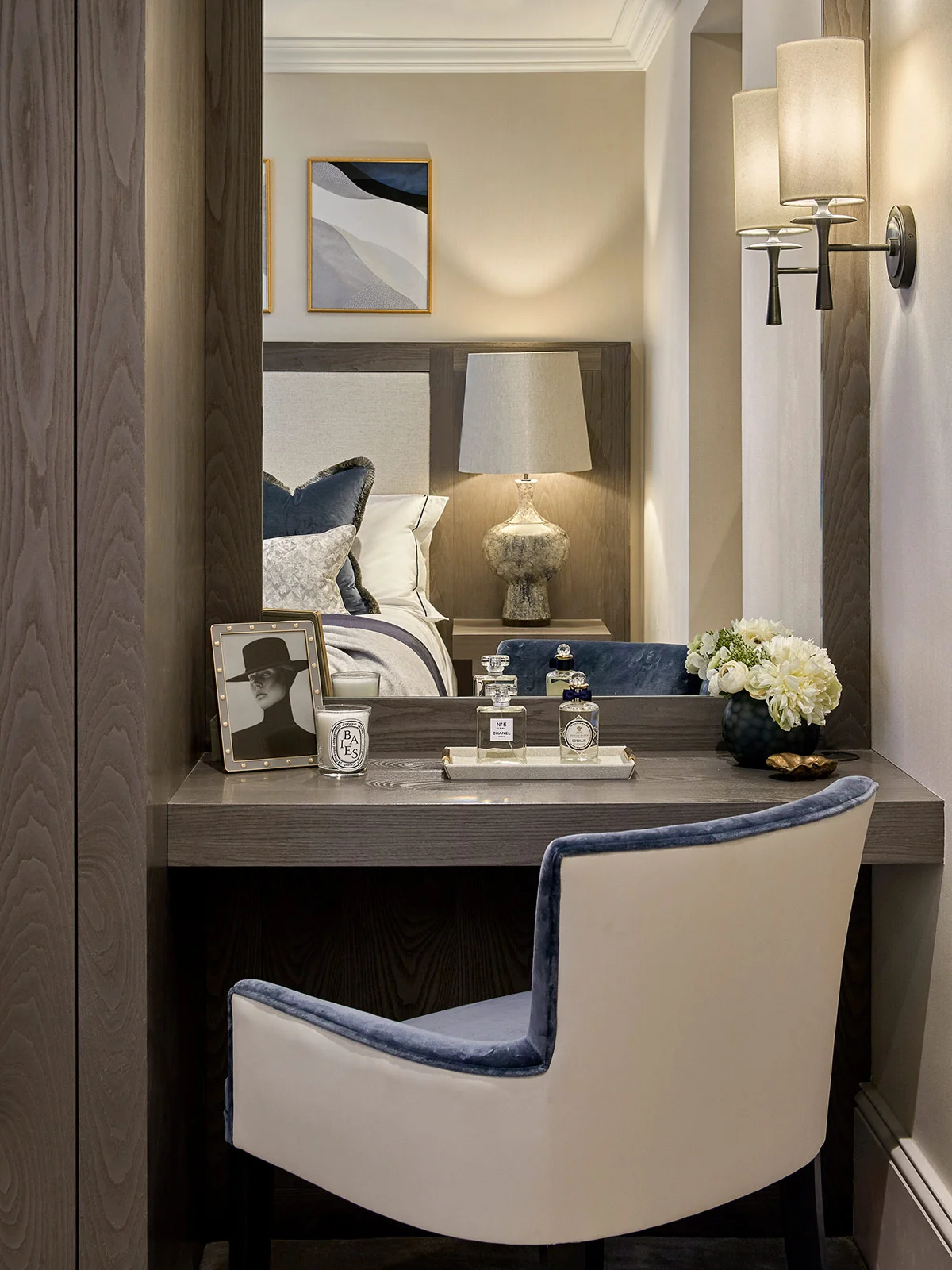 A modern vanity area with a mirror, decorative lamp, a vase of flowers, and a stylish chair.