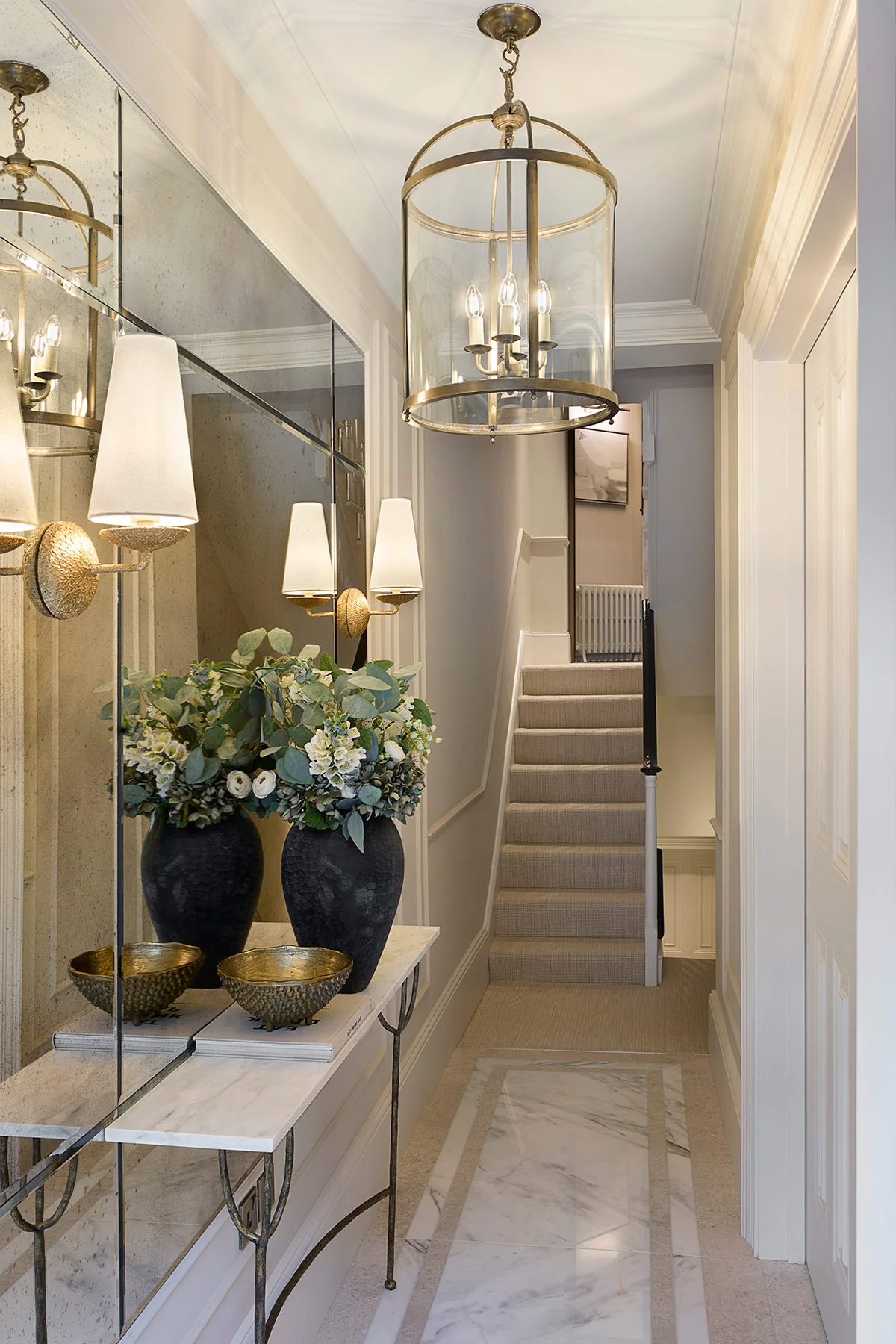 Bright hallway with a staircase, featuring a mirror, two wall sconces, a marble table, and two elegant vases of flowers.