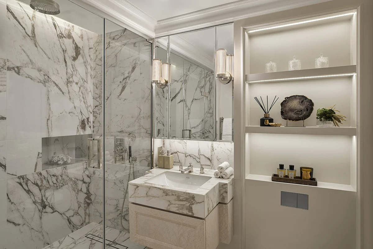 Modern bathroom featuring marble walls, a sleek sink, and stylish lighting. Shelves display decorative items and toiletries.