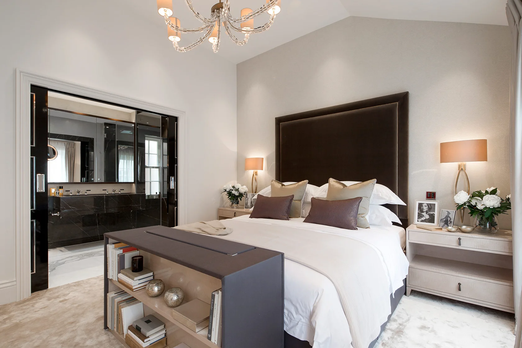 Elegant bedroom featuring a chandelier, plush bedding, and stylish decor, with an ensuite visible in the background.