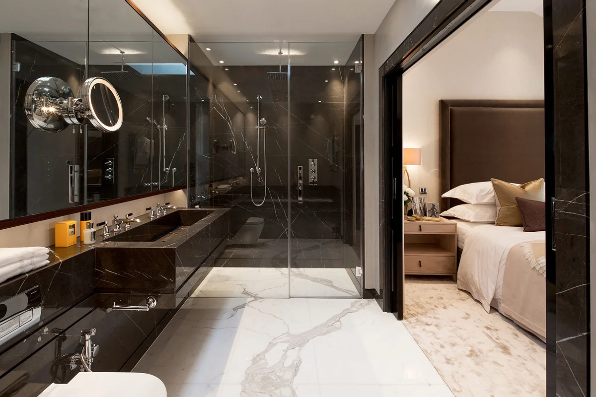 Modern bathroom with glass shower, double sink vanity, and a view into a cozy bedroom with warm lighting.