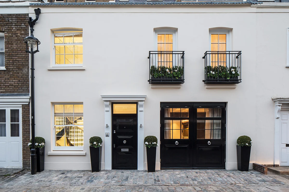 Charming mews house with black doors, balcony, and glowing windows, surrounded by potted plants on a cobbled street.