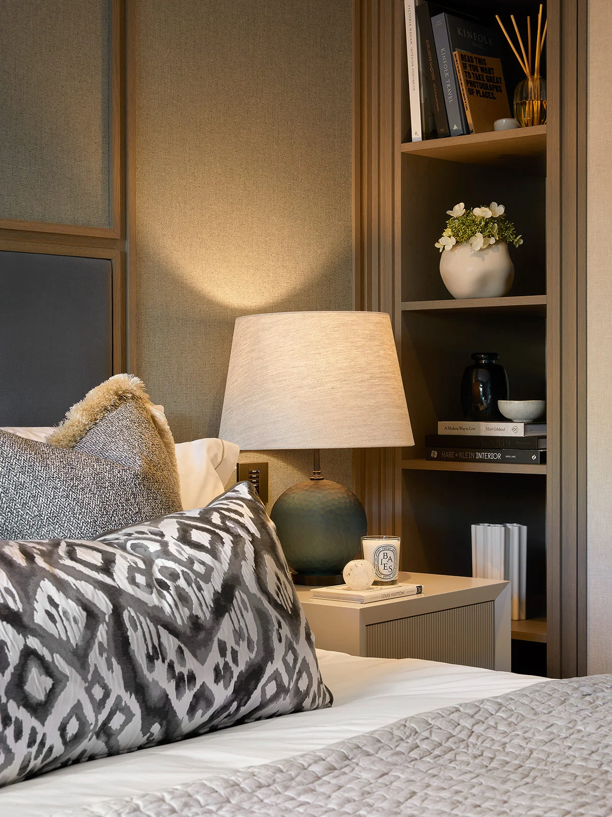 Stylish bedroom corner featuring a cozy bed, decorative pillows, a nightstand with a lamp, and a shelf with decor items.