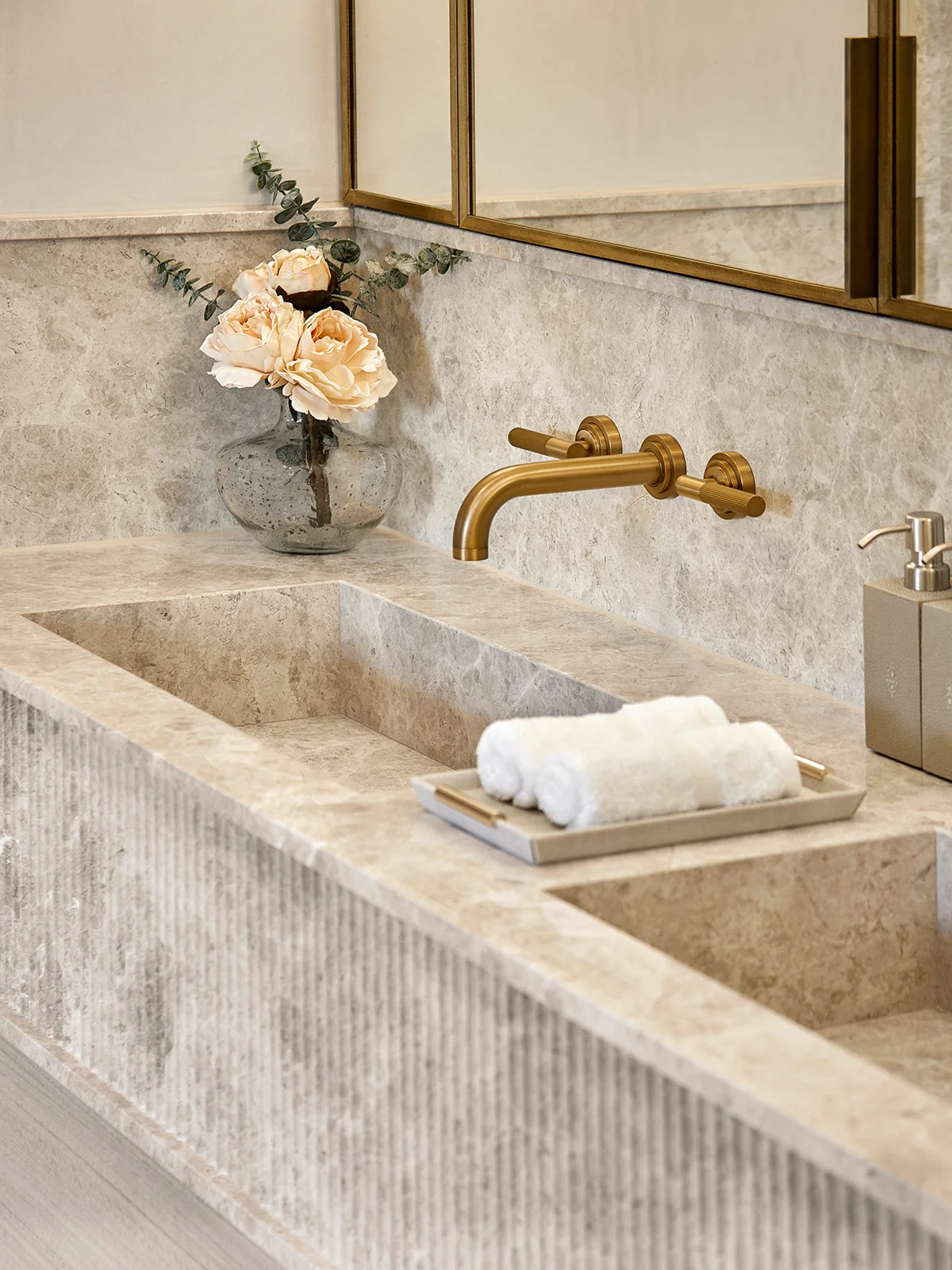 Elegant bathroom sink with gold faucet, white towels on a tray, and a floral arrangement in a glass vase.