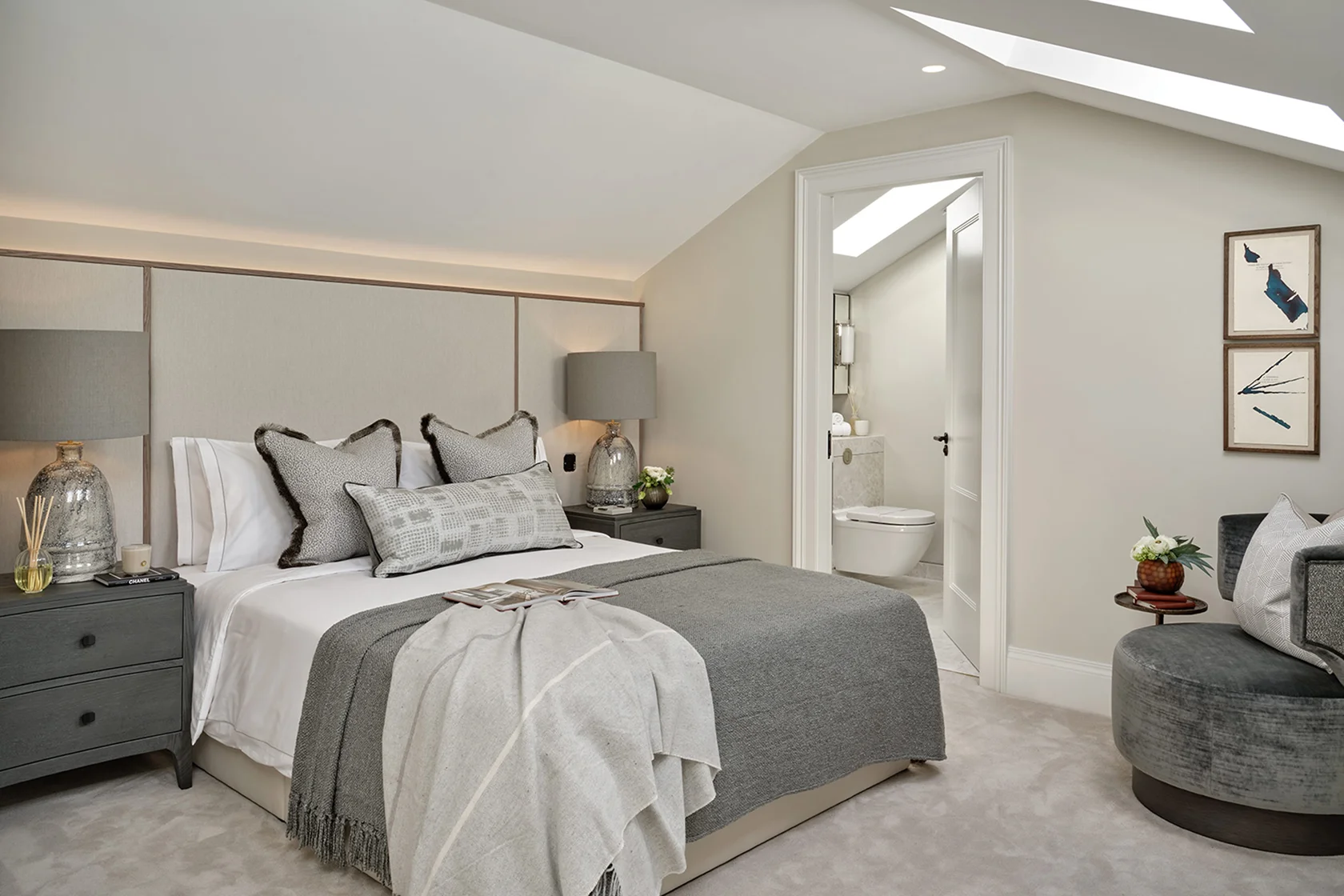 Modern bedroom featuring a gray bedspread, stylish lamps, and an adjoining bathroom visible in the background.