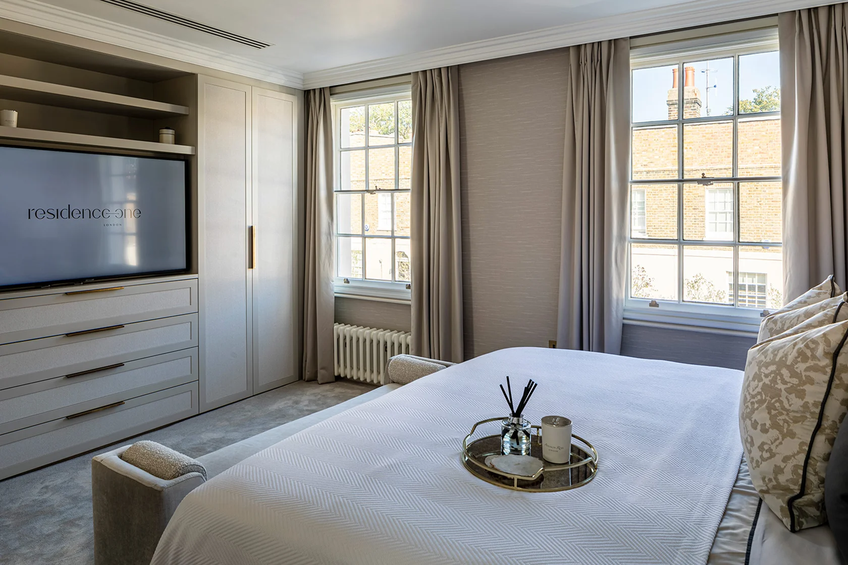 Modern bedroom featuring a large bed, stylish decor, and windows with curtains, alongside a TV and dresser.