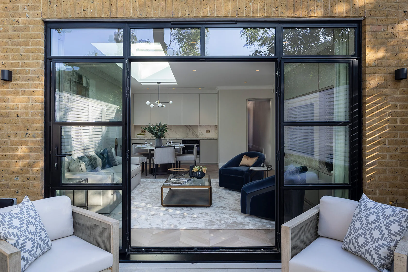 Modern living space viewed through large black-framed glass doors, with outdoor seating in front.