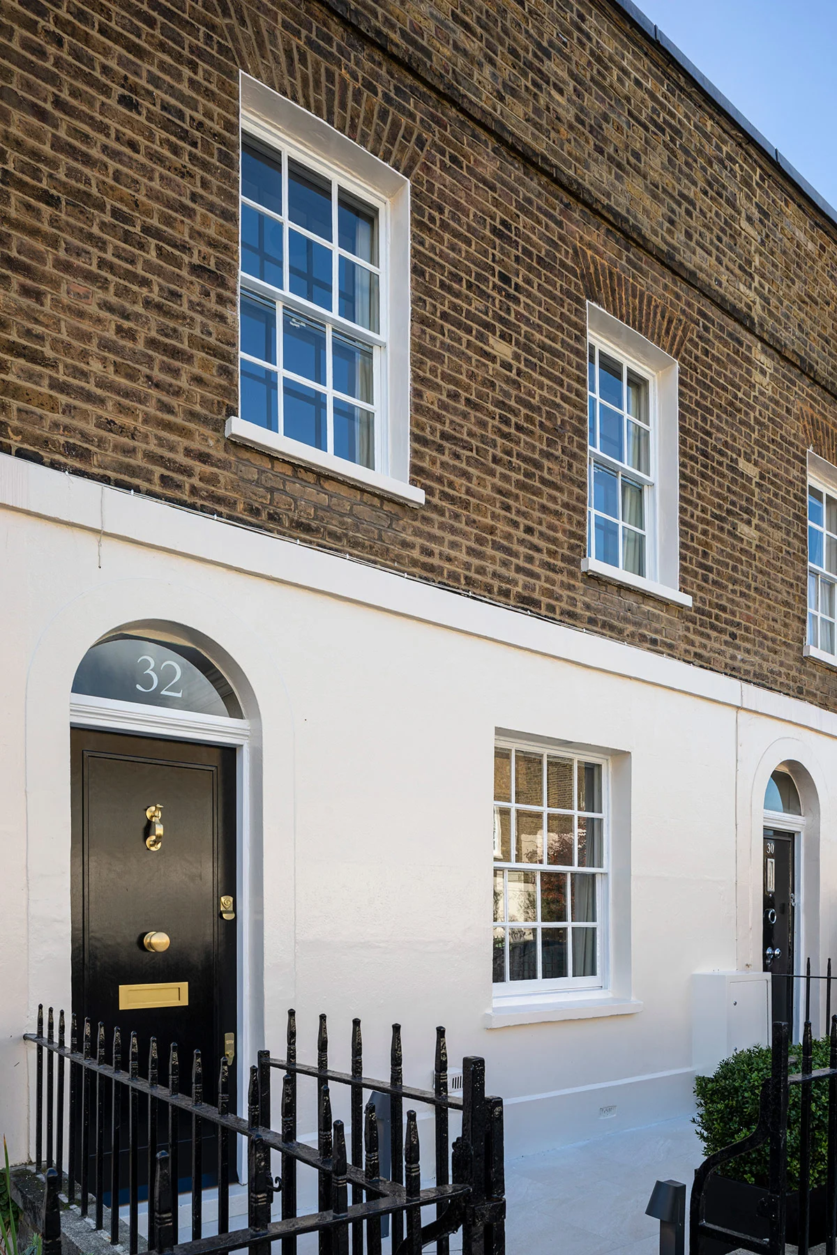 Exterior view of a brick building with black door and white window frames, bordered by a small iron fence.