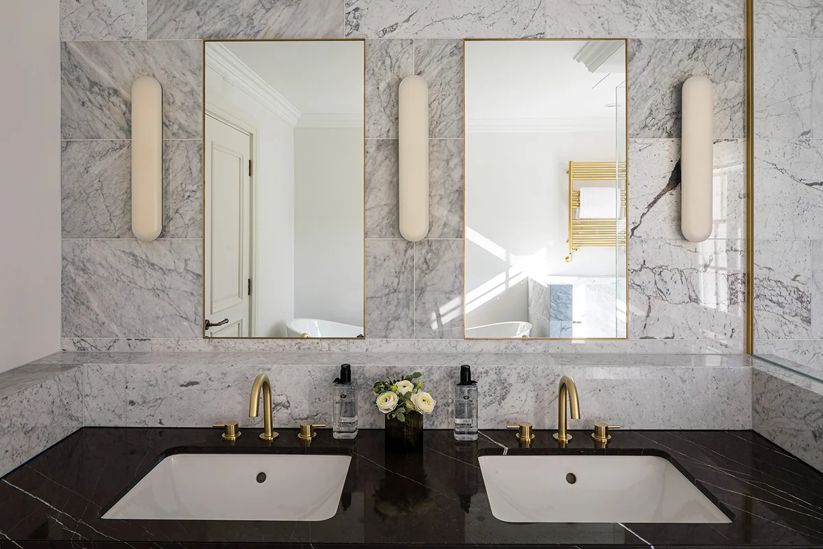 Modern bathroom with dual sinks, marble walls, and elegant mirrors, featuring soft lighting and decorative accessories.