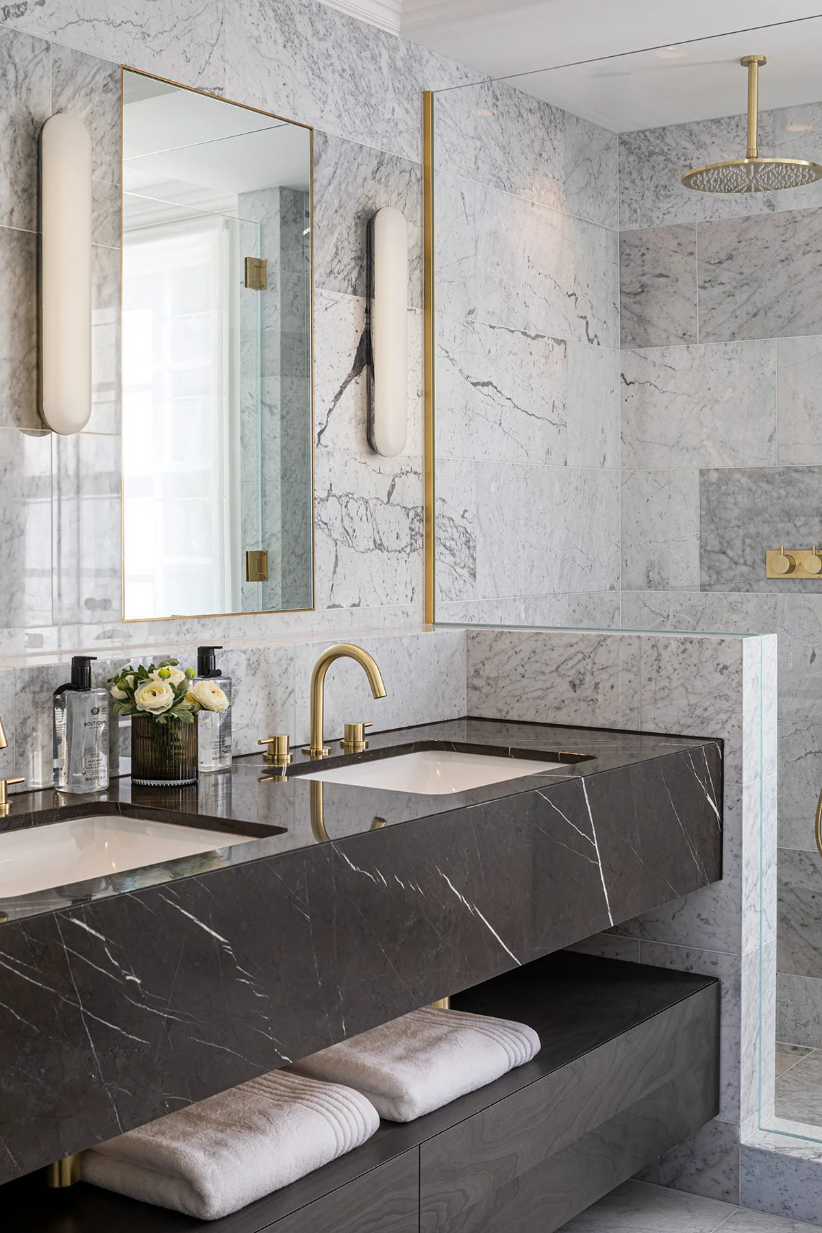 Modern bathroom featuring marble walls, dual sinks with gold fixtures, and a glass shower enclosure.