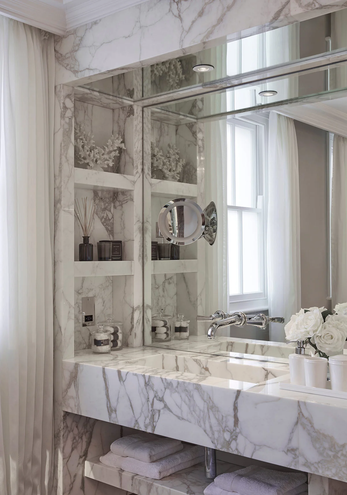 Luxurious bathroom with marble walls, a large mirror, shelves with toiletries, and white flowers.