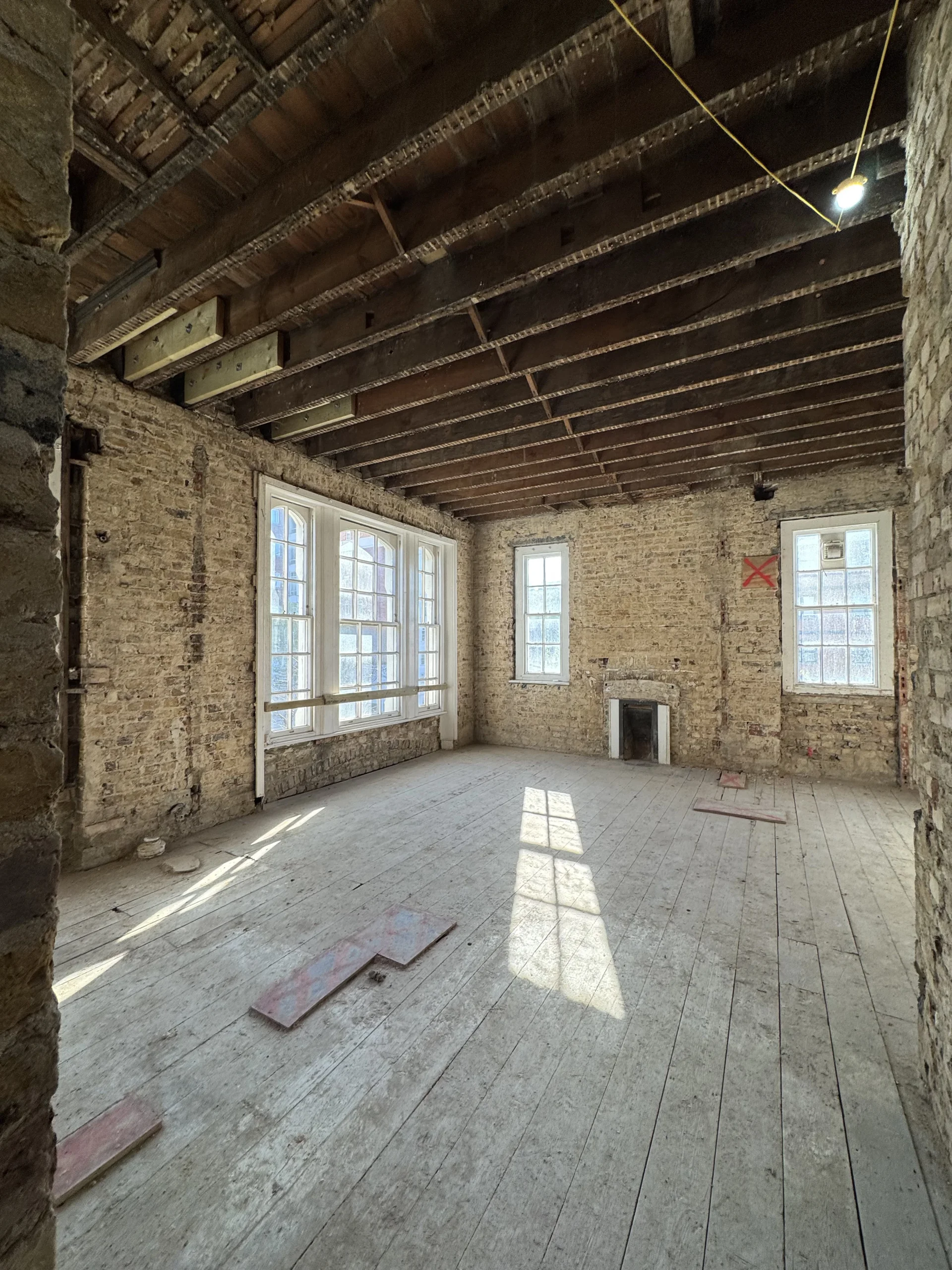 Renovated room with exposed brick walls, large windows, wooden beams, and a wooden floor illuminated by natural light.